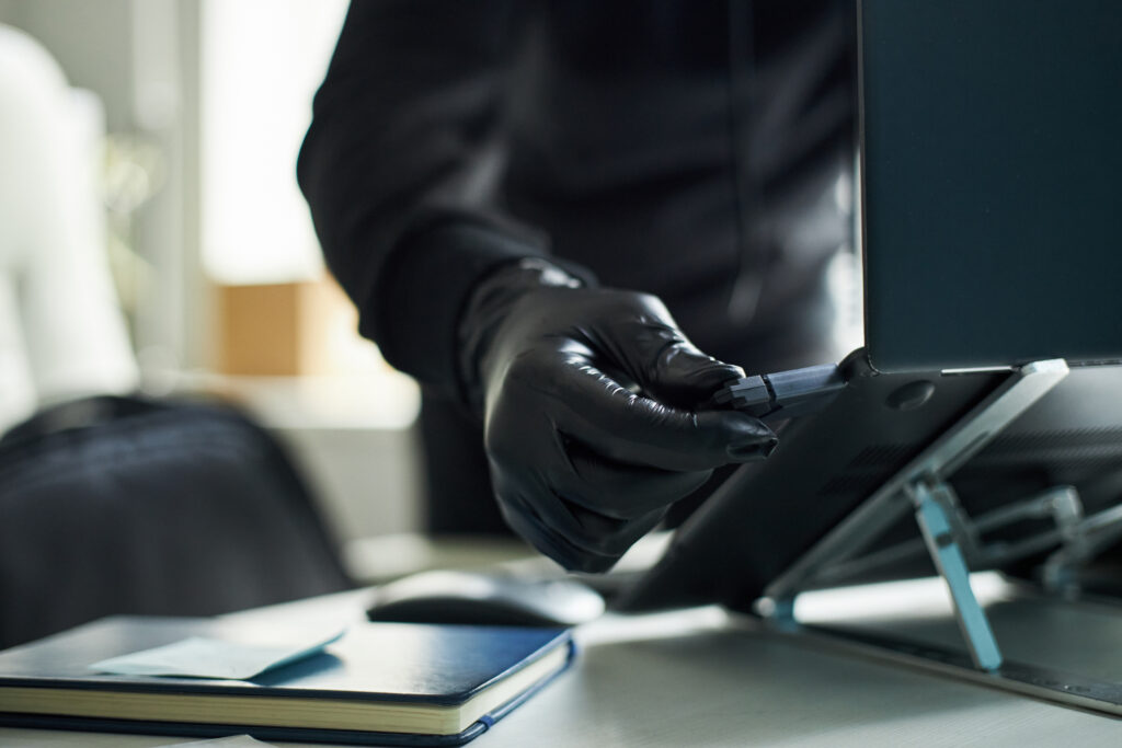 A person wears gloves, inserting a USB device into a laptop. A notebook and papers sit on the desk in a bright room.