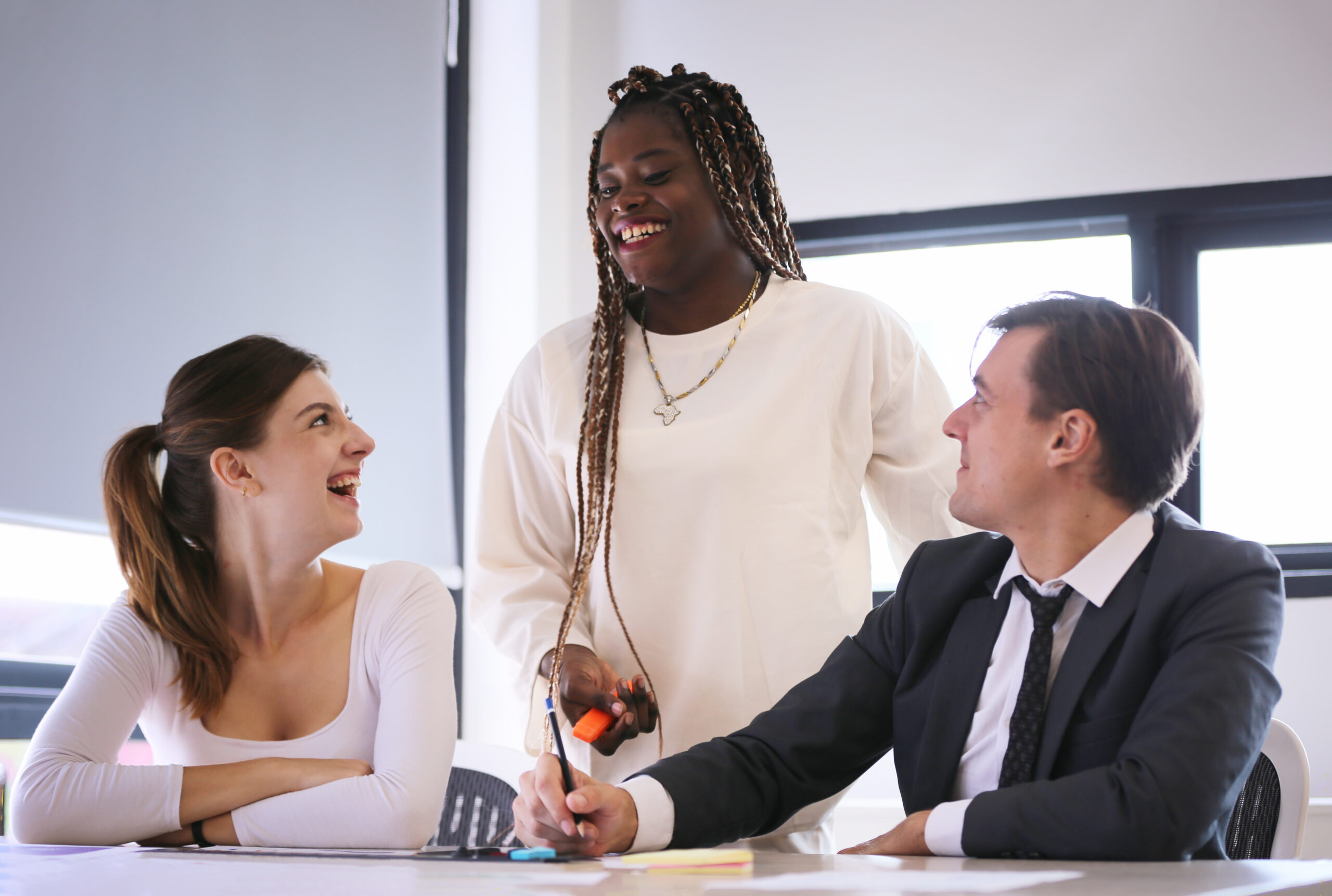 Three people in an office setting, interacting and smiling around a desk with papers and pens, in front of large windows.