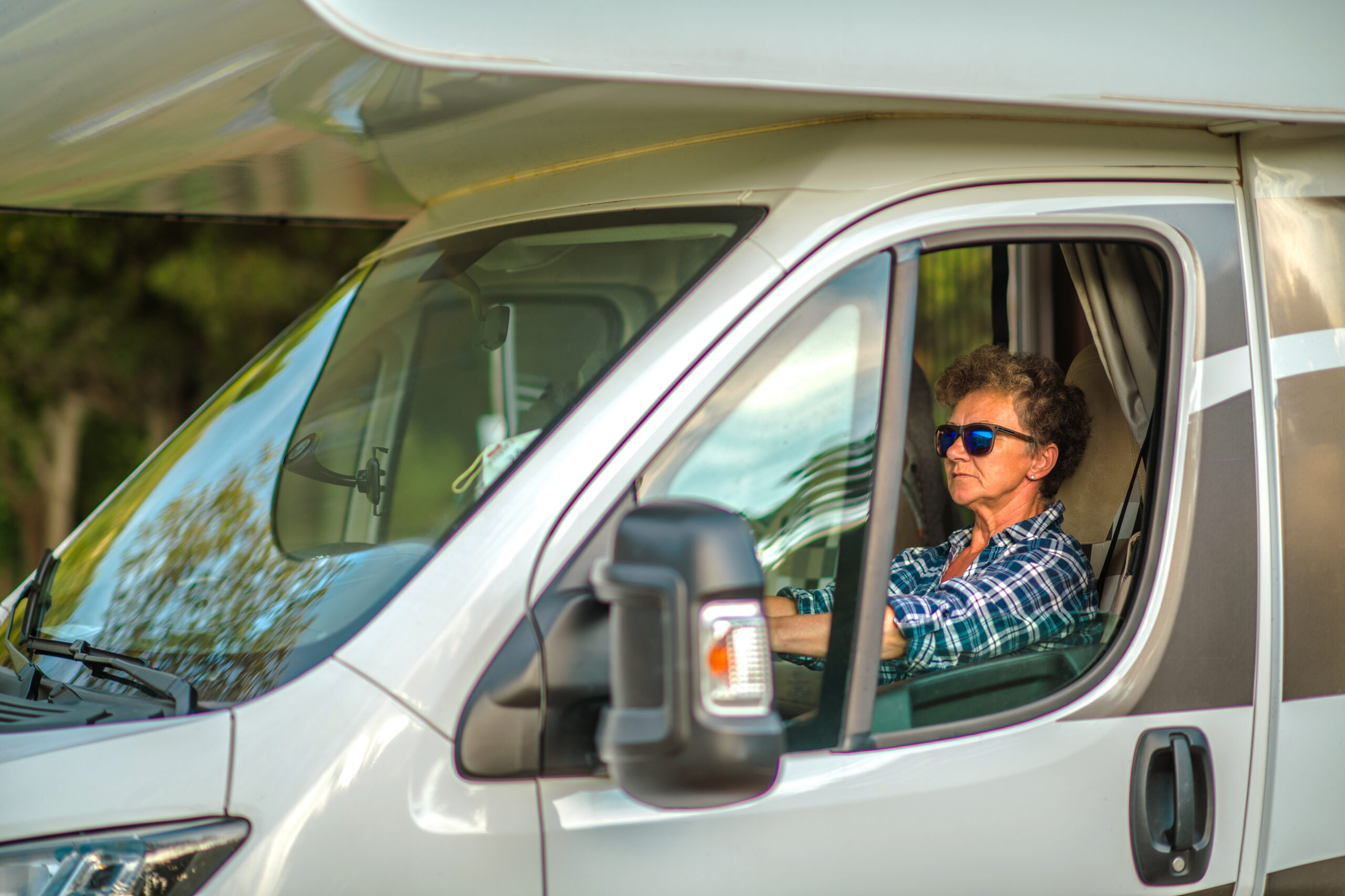 A person wearing sunglasses drives a white camper van. The vehicle is parked near trees, suggesting a serene outdoor setting.
